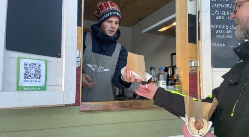Ung man i förkläder sträcker fram en korv med bröd till en kund.