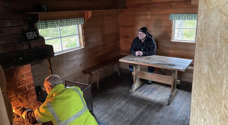 En man sitter vid ett helt bord och tittar på när en man i reflexväst tänder en brasa i öppna spisen.