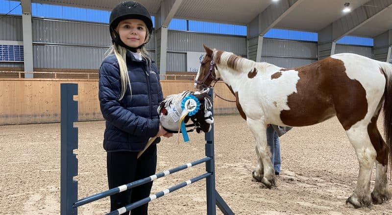 Flicka med ridhjälm och käpphäst framför ett hinder inne i ridhuset. I bakgrunden står en häst som liknar käpphästen