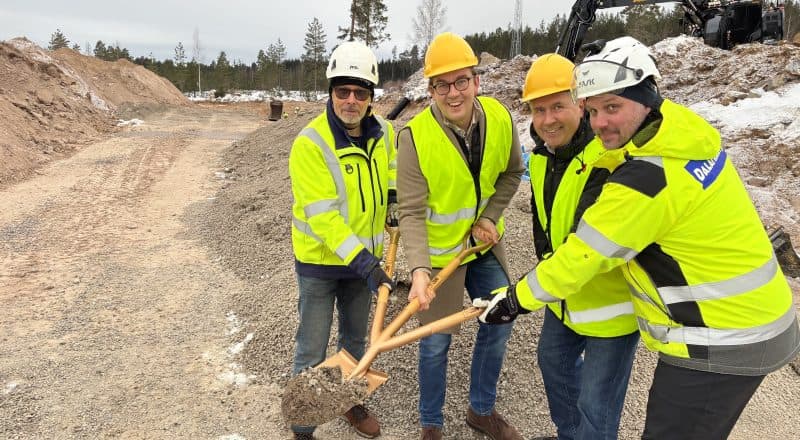 Fyra personer håller i en spade med grus på bladet och tre skaft.