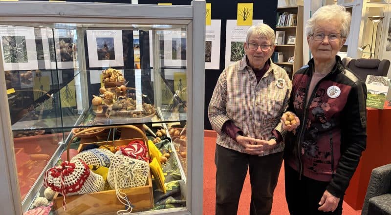 Två kvinnor står och håller i lökar med en glasmonter intill. I bakgrunden syns utställningens affischer.