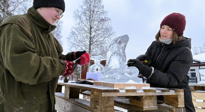 Två elever karvar i ett isblock som ser ut som en fisk eller en ål.