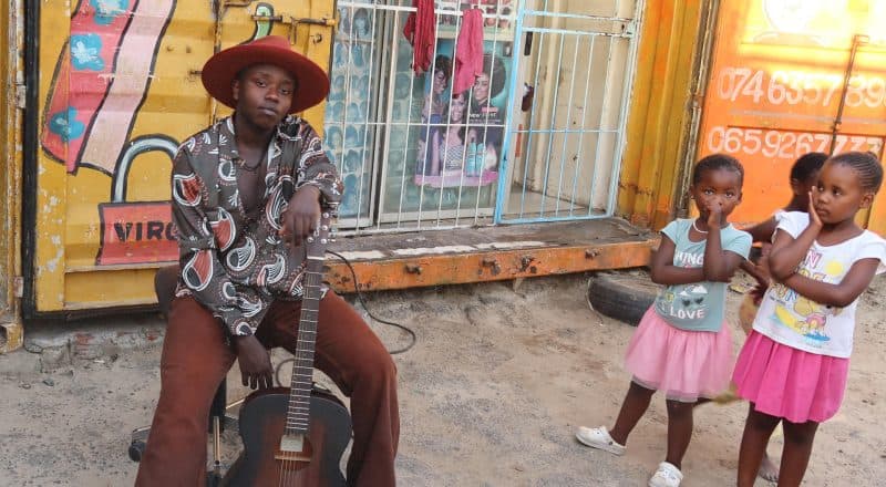 Toussaint "Tusse" Chiza poserar med en gitarr på en afrikansk gata med barn i bakgrunden.