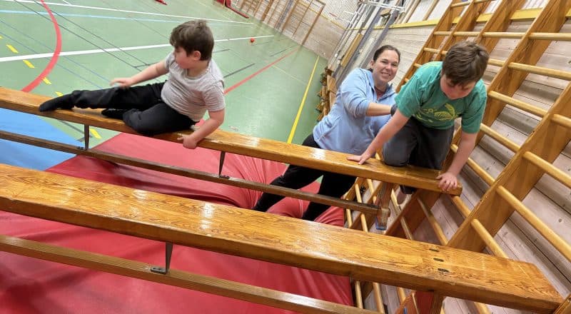 Närbild på barn som klättrar upp för en ribbstol och glider ned längs en bänk. Mamman Nikolett hjälper till i bakgrunden.