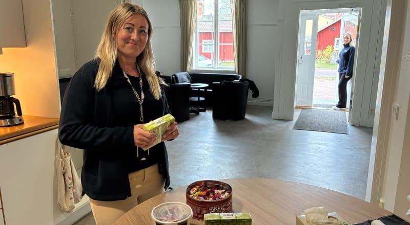 En kvinna förbereder ett bord med fika och dryck, en annan kvinna håller upp en dörr in till lokalen.