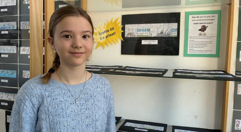 Mellanstadieelev uppställd framför glasskåp där teckningen hänger på väggen tillsammans med en "Grattis"-skylt.