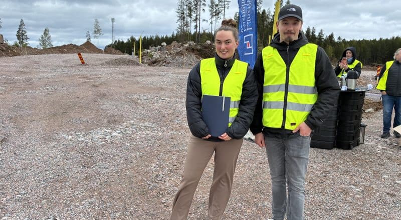 Två personer i varselvästar och med ett mäklarprospekt i famnen tittar in i kameran.
