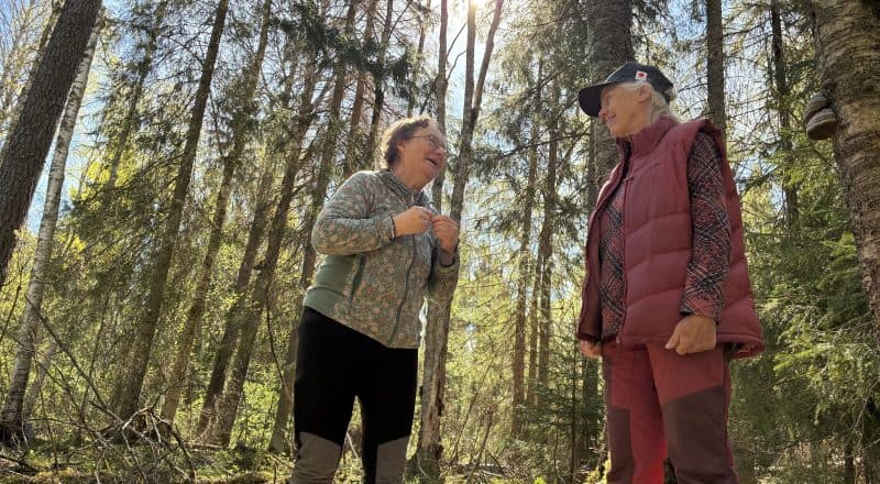 Två kvinnor står i en grönskande skog och pratar och skrattar med varandra.