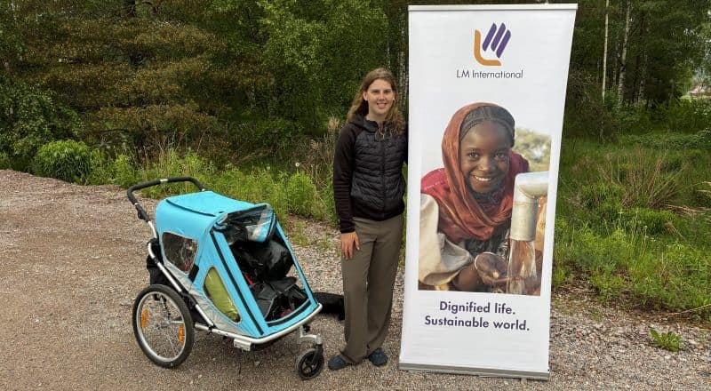 Ung kvinna står mellan löparbarnvagn och en stor affisch med en kvinna som hämtar leende afrikansk kvinna som vatten ur ett rör.