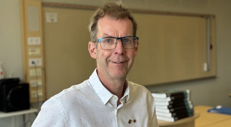 Lars Halvarson ler i ett klassrum, iklädd vit skjorta med skjortnålar med Leksands kommunvapen och Socialdemokraternas ros.
