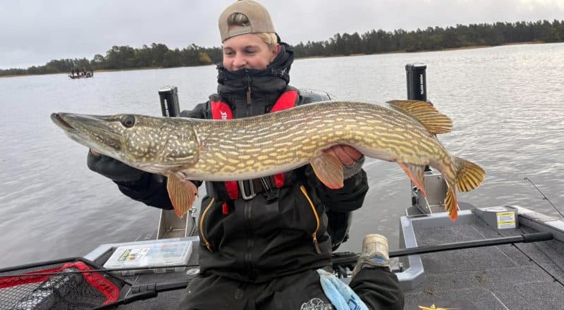 Max Persson sitter i en båt med regnkläder och flytväst, framför sig håller han en meterlång gädda.