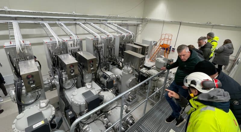 Ett ställverk, en stor metallisk apparat med ledningar och rör, fotograferat från en läktare med personer som står och pekar på aggregatet.