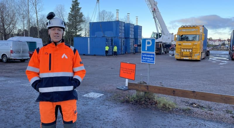 Man i varselkläder och hjälm framför en lyftkran som lyfter containrar på plats.