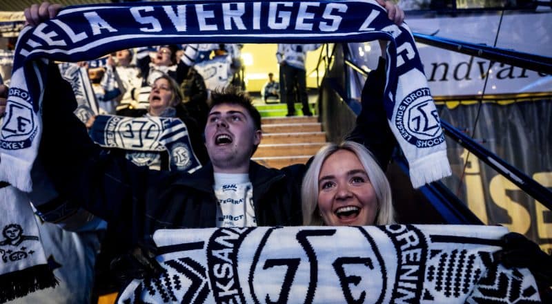 Två personer i Leksandshalsdukar hejar engagerat på läktaren.