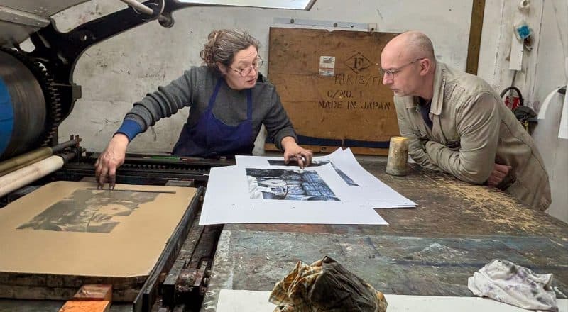 Två personer står lutade över ett bord med en målning på ett papper. Till vänster syns den stora tryckpressen.