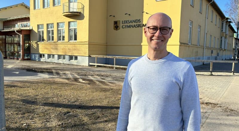 Rektorn står i en långärmad tröja och ler framför en solbelyst skolbyggnad.