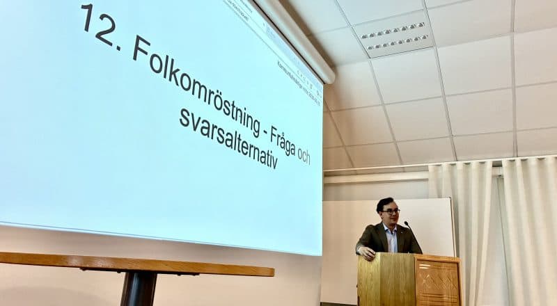 Man står i talarstolen, på skärmen till vänster står "12. Folkomröstning - fråga och svarsalternativ".