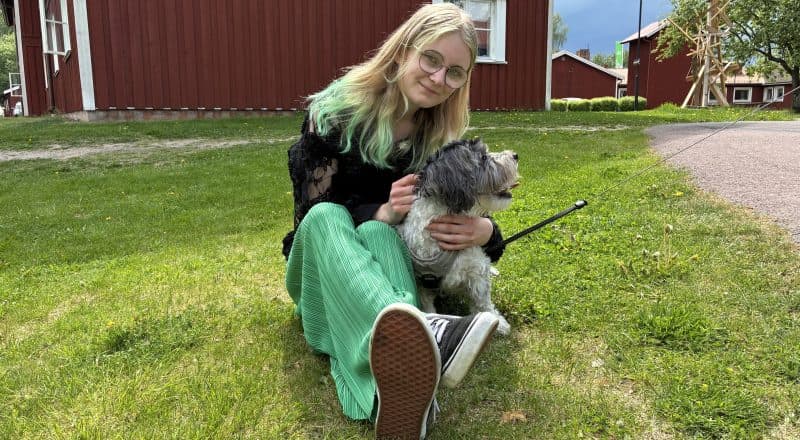 Ung kvinna med gröna byxor och gröna slingor i håret sitter i gräset och klappar en liten lurvig hund.