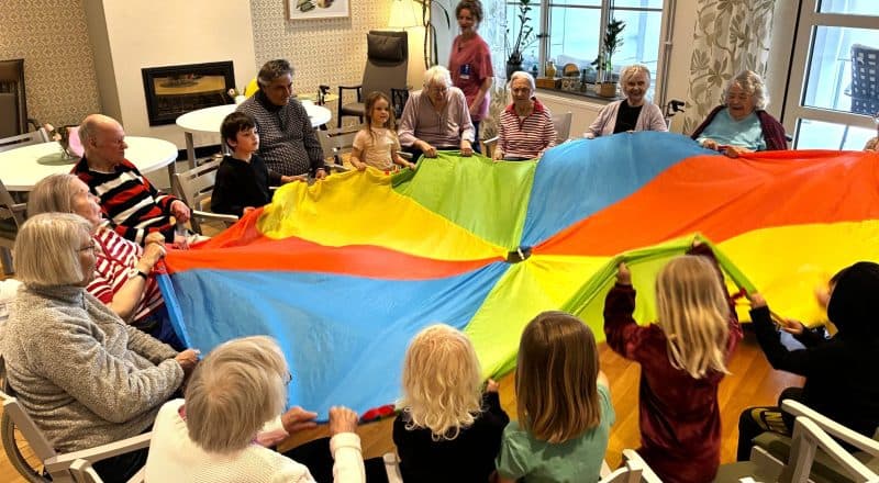 Ungefär 25 deltagare i en ring håller i ett färgglatt fallskärmsnät som en boll rullar runt på.