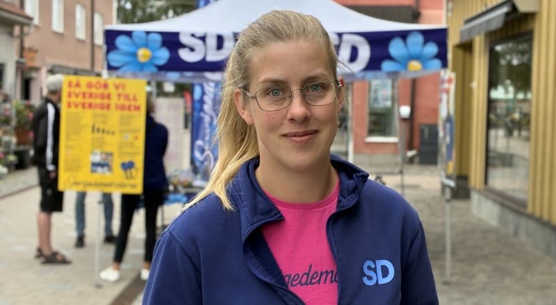 Kvinna i SD:s partikläder med blå jacka och rosa tröja framför ett valtält med SD:s symboler på gågatan i Leksand.