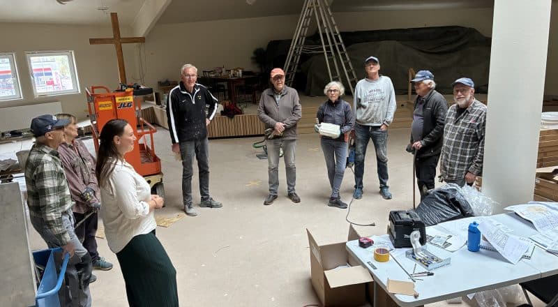 Nio personer uppställda i en halvcirkel på en byggarbetsplats inomhus.