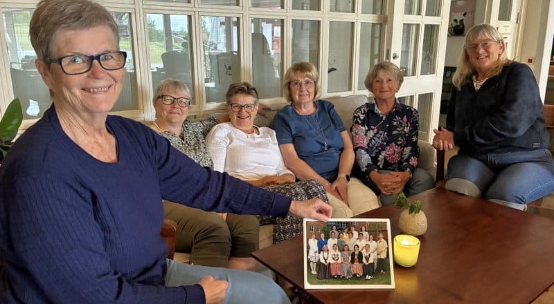 Sex damer sitter i en soffa i en hotell-lobby. Kvinnan närmast kameran håller upp en gruppbild från när de utbildade sig på 90-talet.
