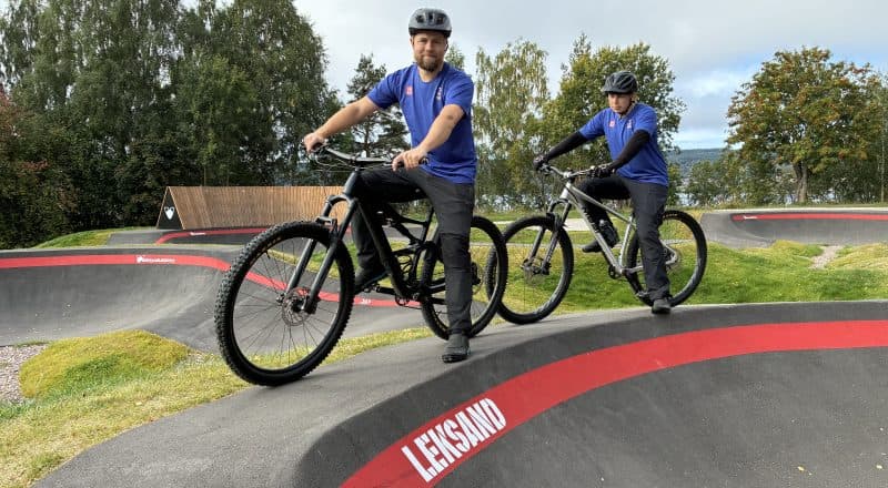 Två vuxna cyklister i blå kläder på varsin cykel vid pumptrackbanans kant.