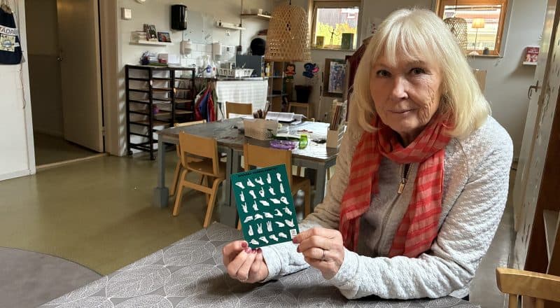 Kvinna med halsduk sitter i ett tomt rum på en förskola och håller upp ett vykort som visar handalfabetet.