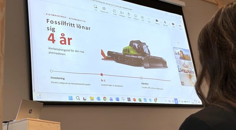 Bild på en presentation som visas på storbildsskärm, där en eldriven pistmaskin visas upp med detaljer som att fossilfritt lönar sig inom fyra år. En åskådares hår skymtar i förgrunden.
